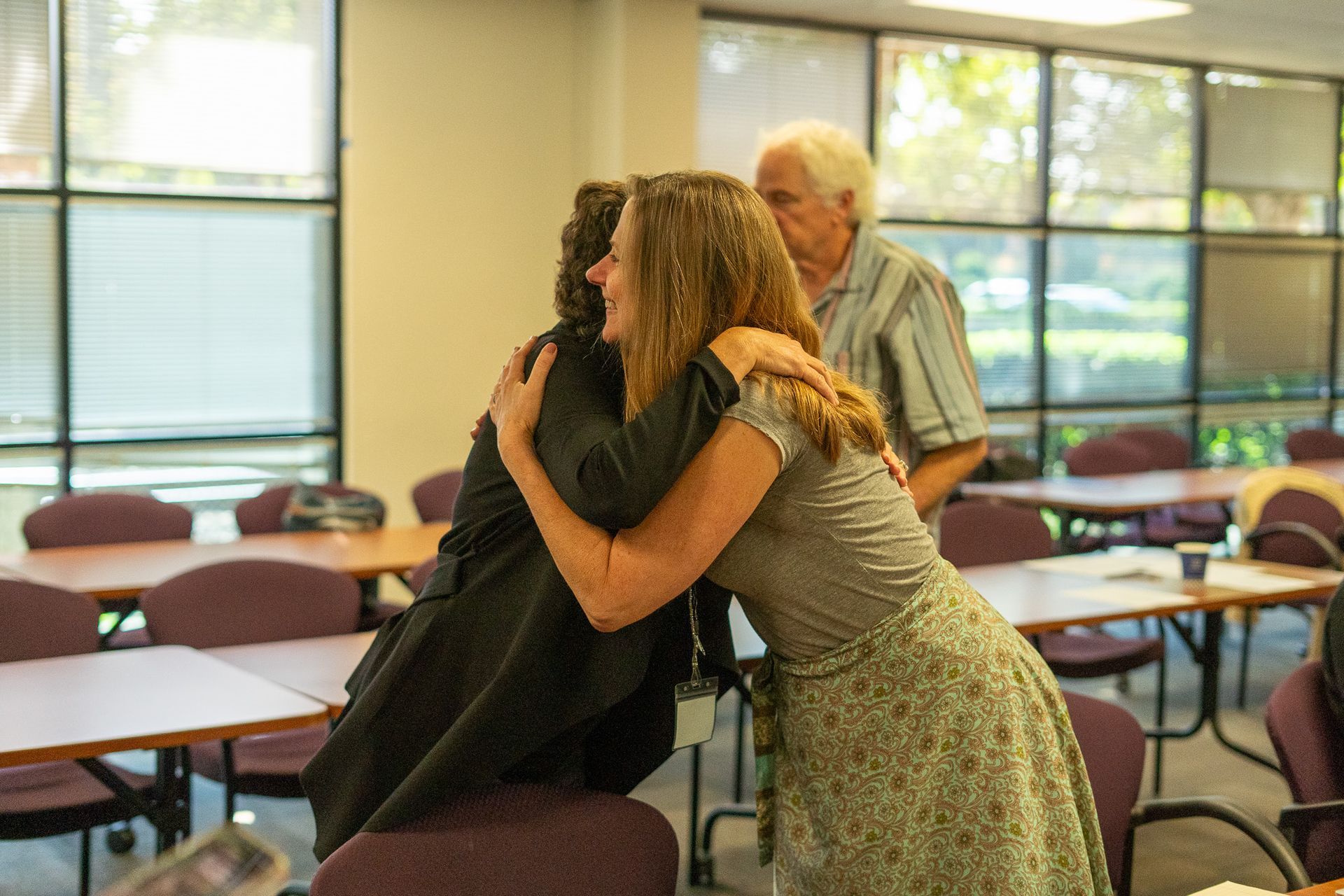 Two people hugging in a classroom, with tables, chairs, and large windows in the background.