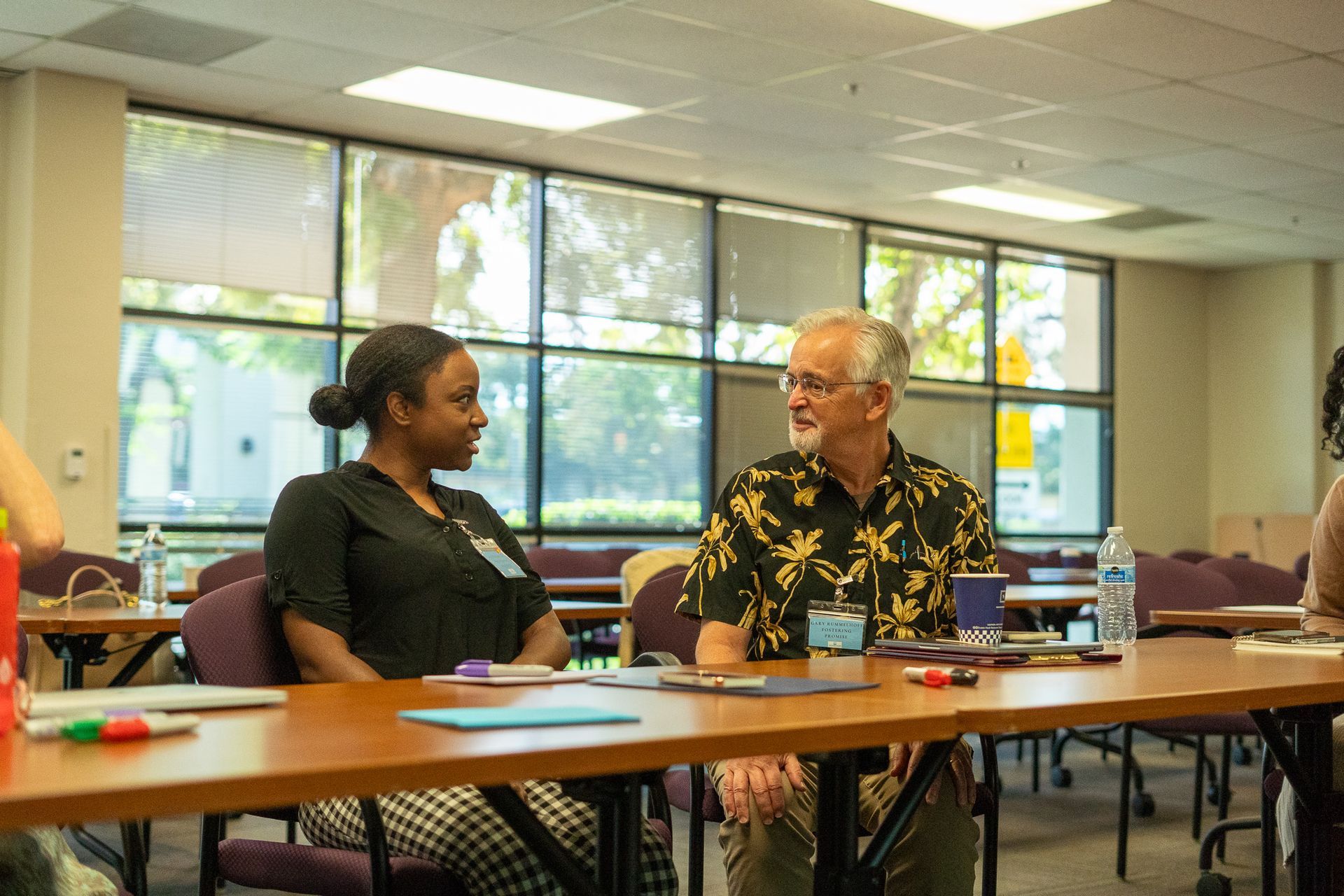 Two people talking at a table in a bright room with large windows and chairs around them