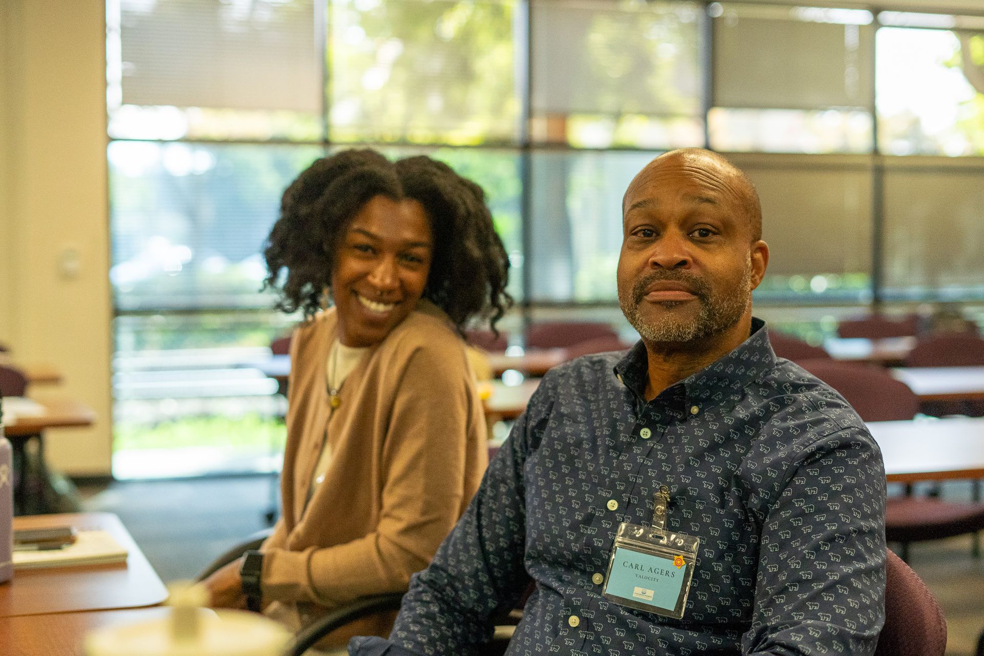Two people sitting at a table in a bright indoor space, smiling at the camera.