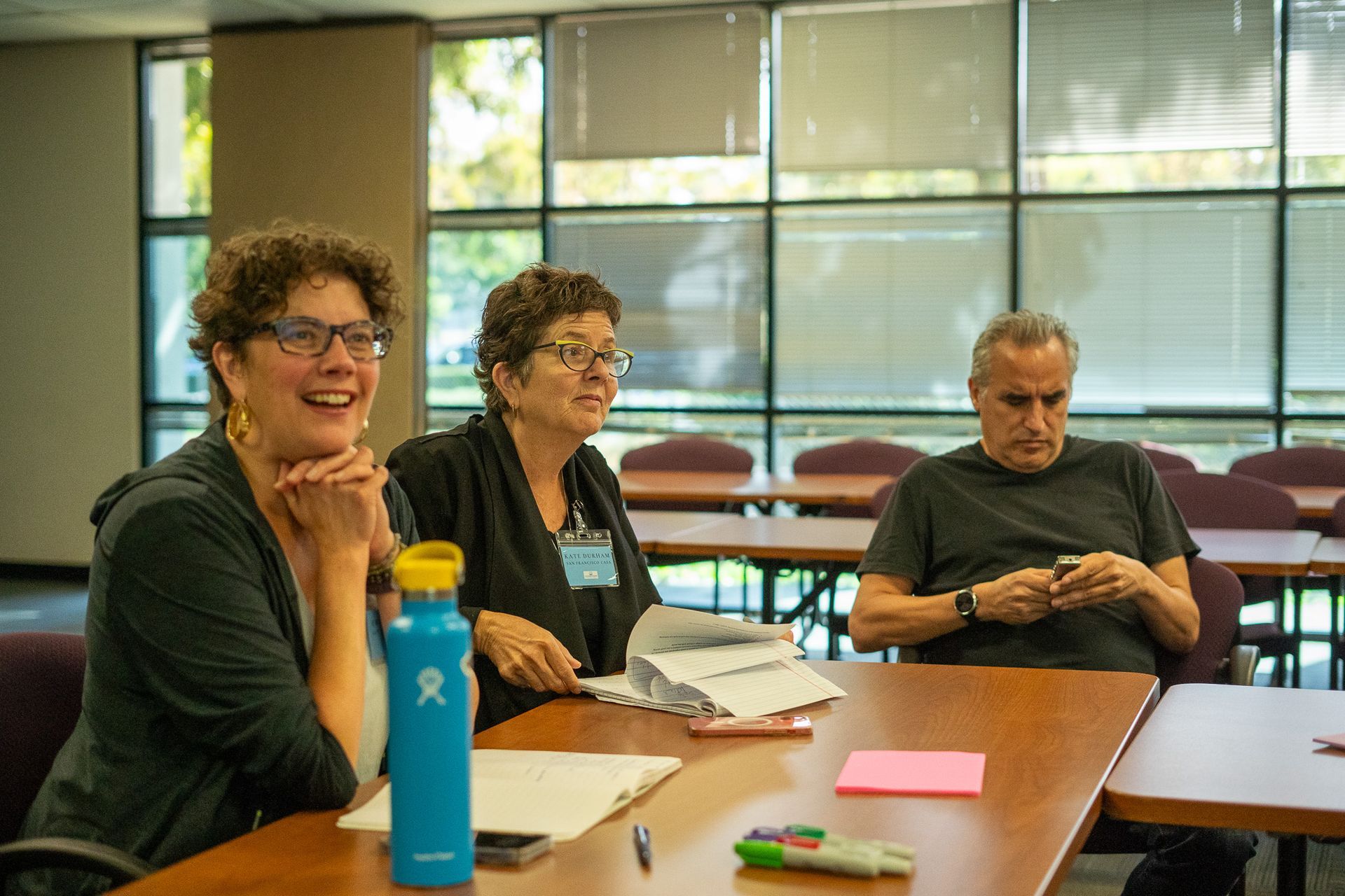 Three people seated at a table in a bright meeting room, with papers, pens, and a blue water bottle.