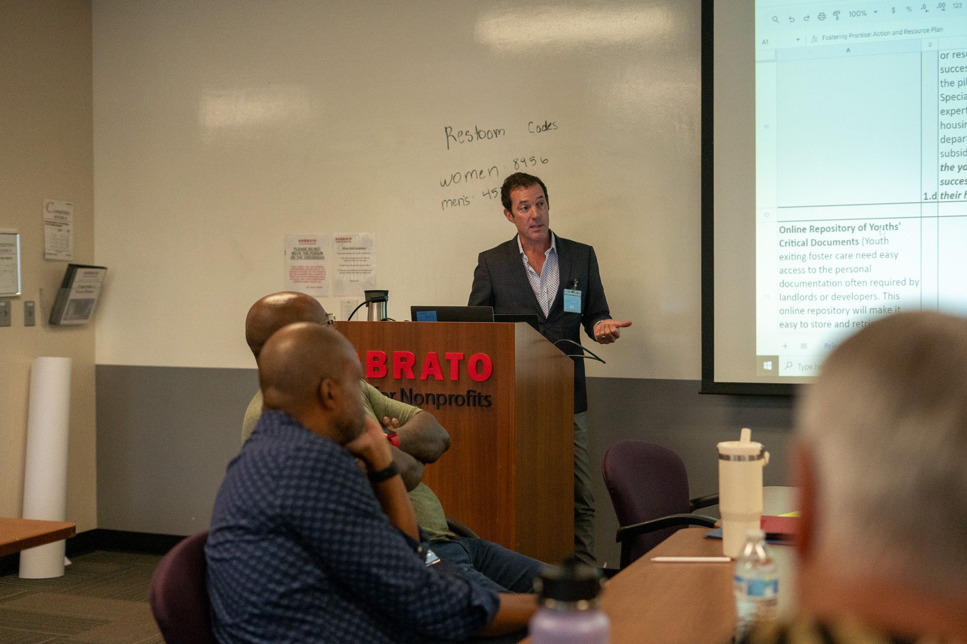 Speaker at podium addressing an audience in a classroom, with a projected screen beside him.