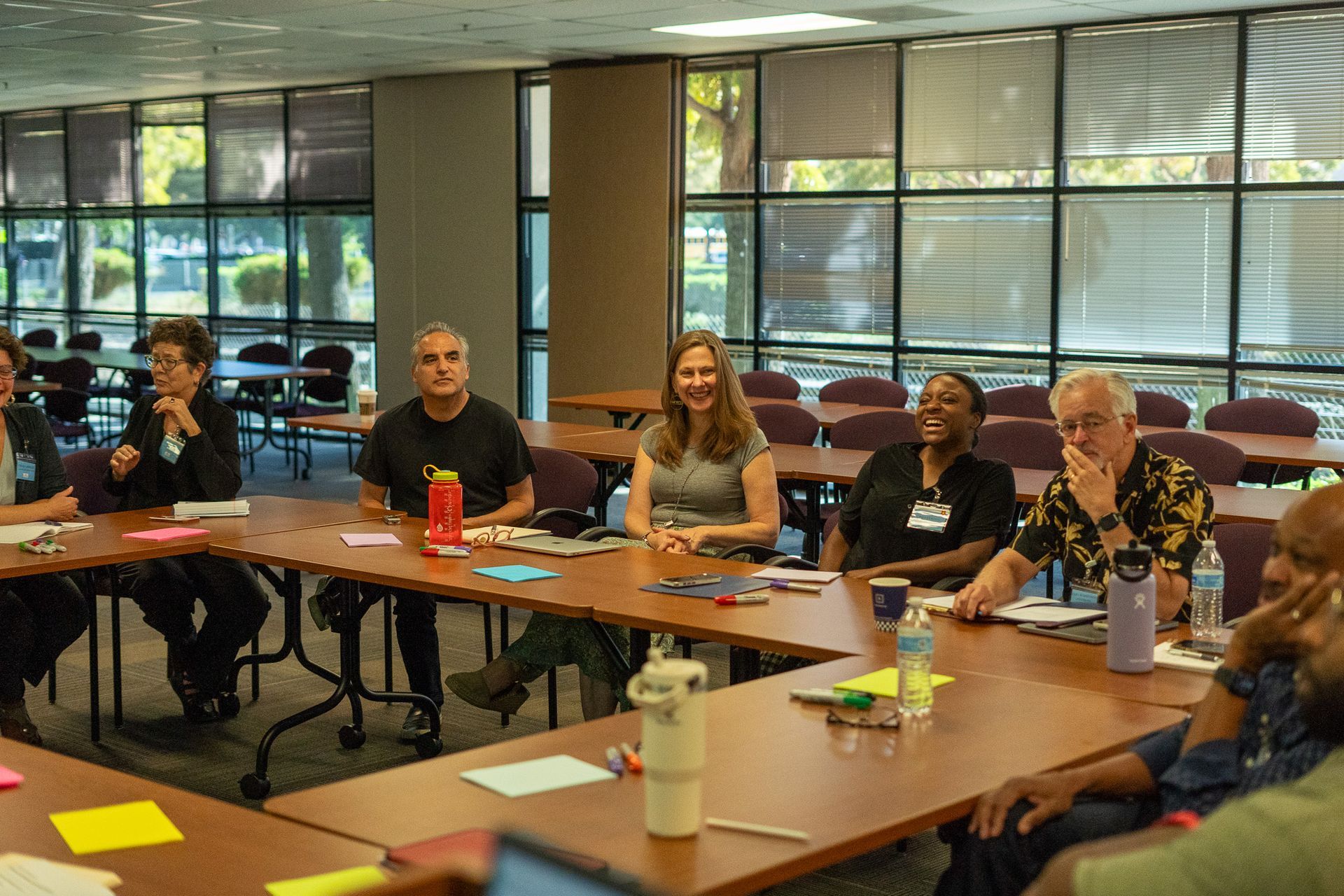 People seated around conference tables in a meeting room with large windows.
