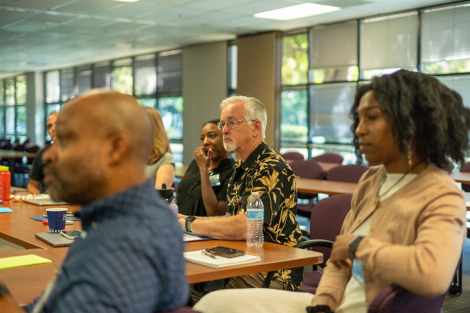 People seated at desks in a classroom or meeting room, listening attentively.