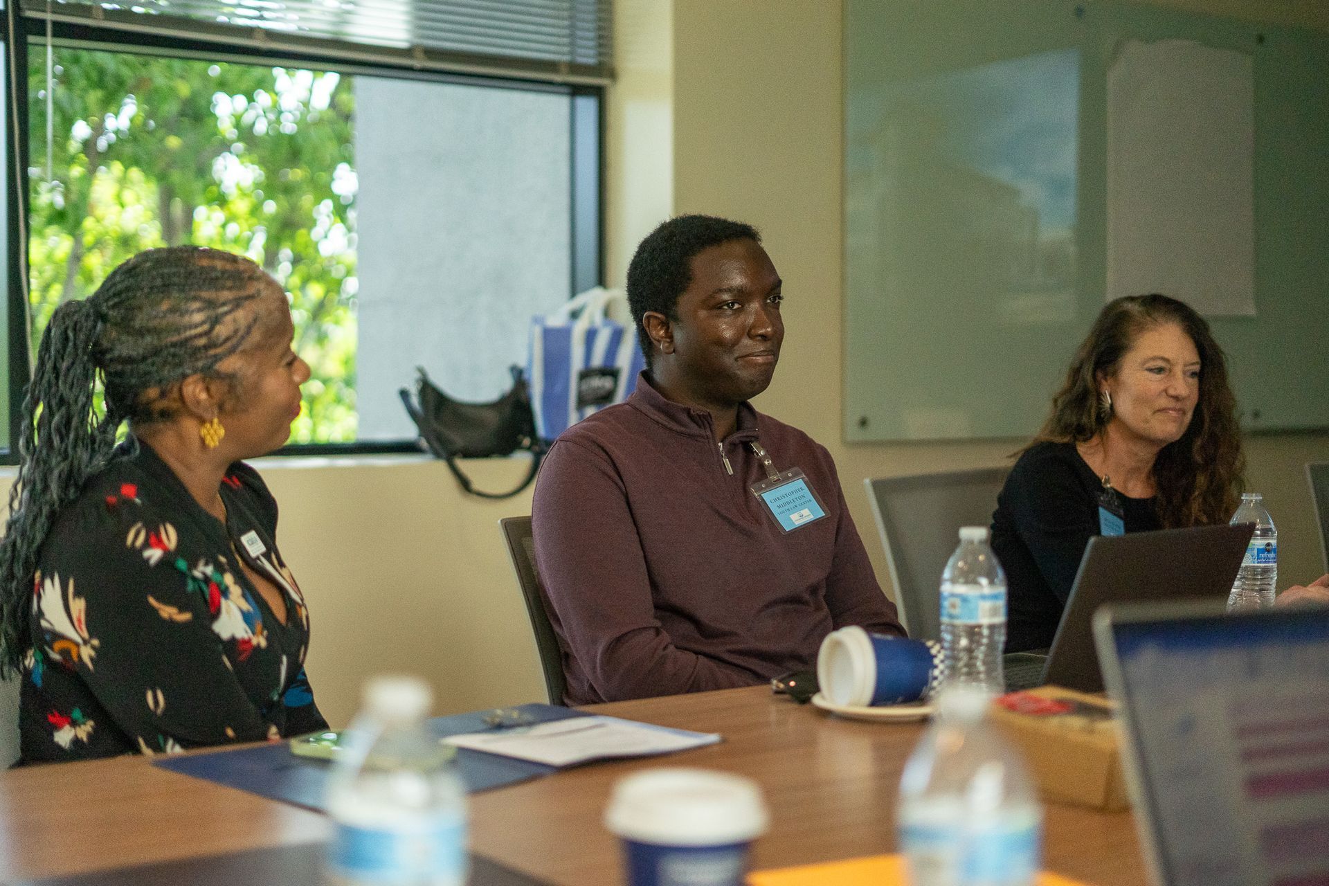 Three people seated at a conference table in a meeting room, with laptops, cups, and papers.