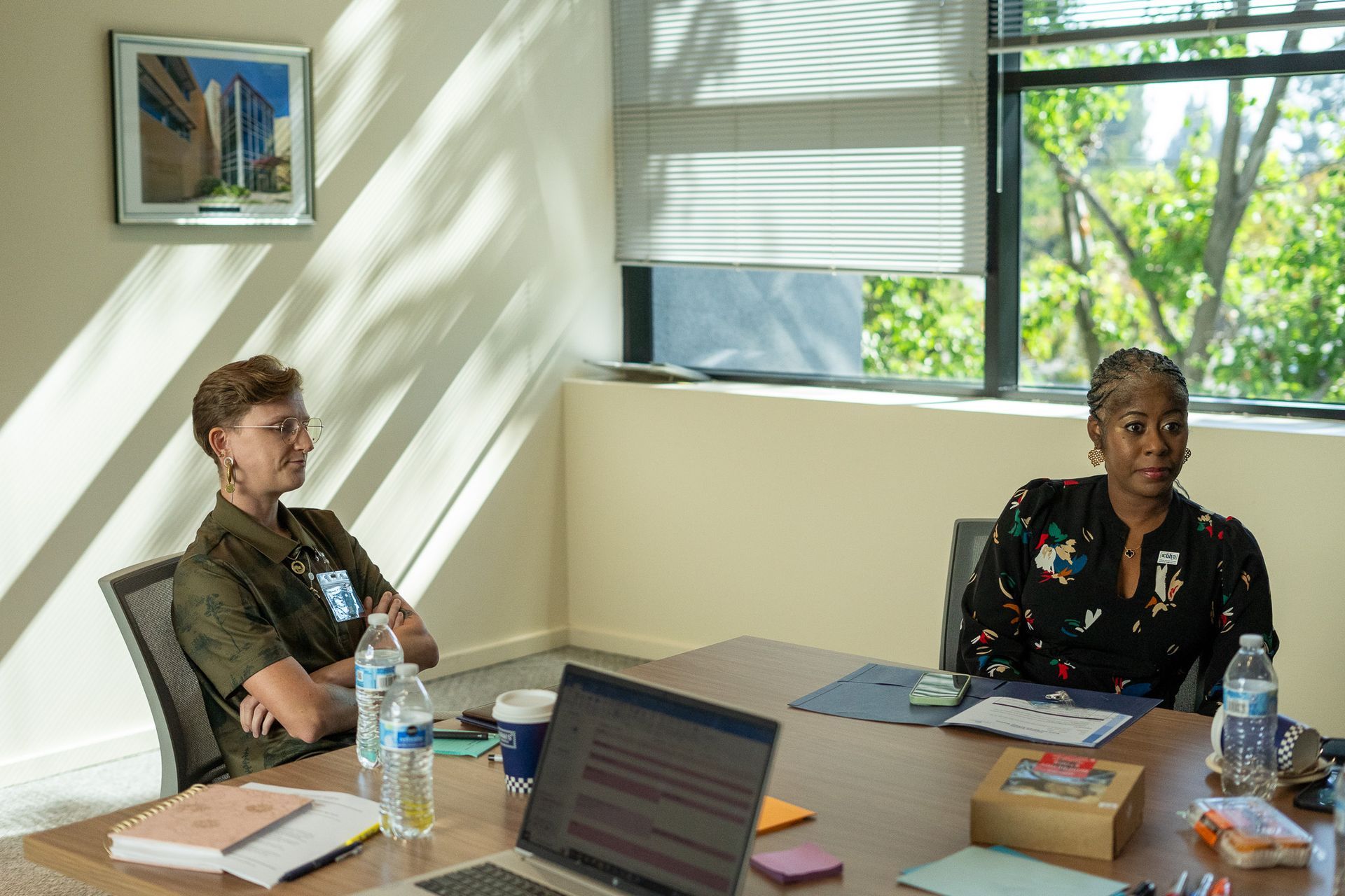Two people seated at a conference table in a bright meeting room with laptops, papers, and water bottles.