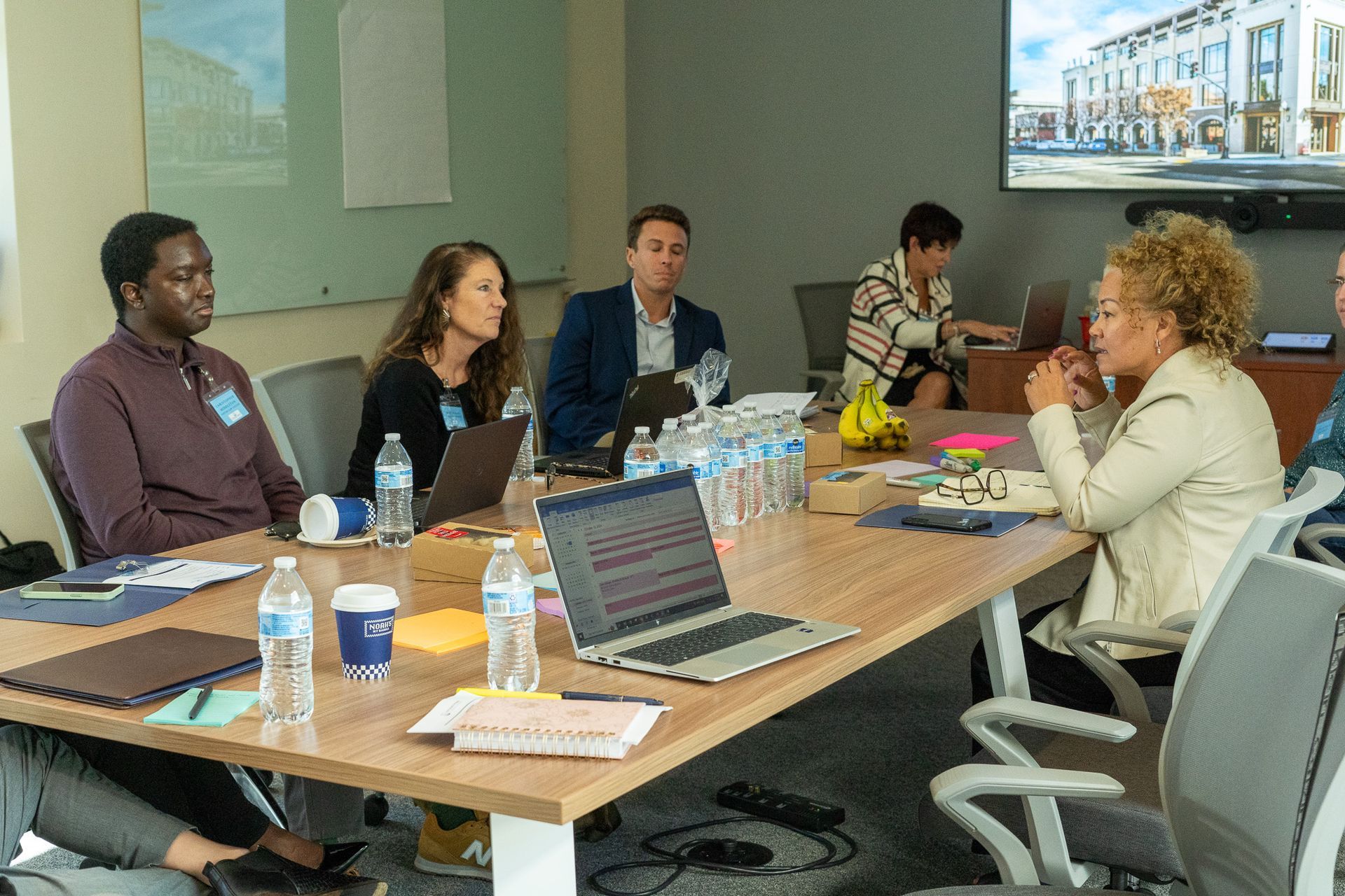 Meeting in a conference room with five people seated around a table with laptops and drinks, discussing together