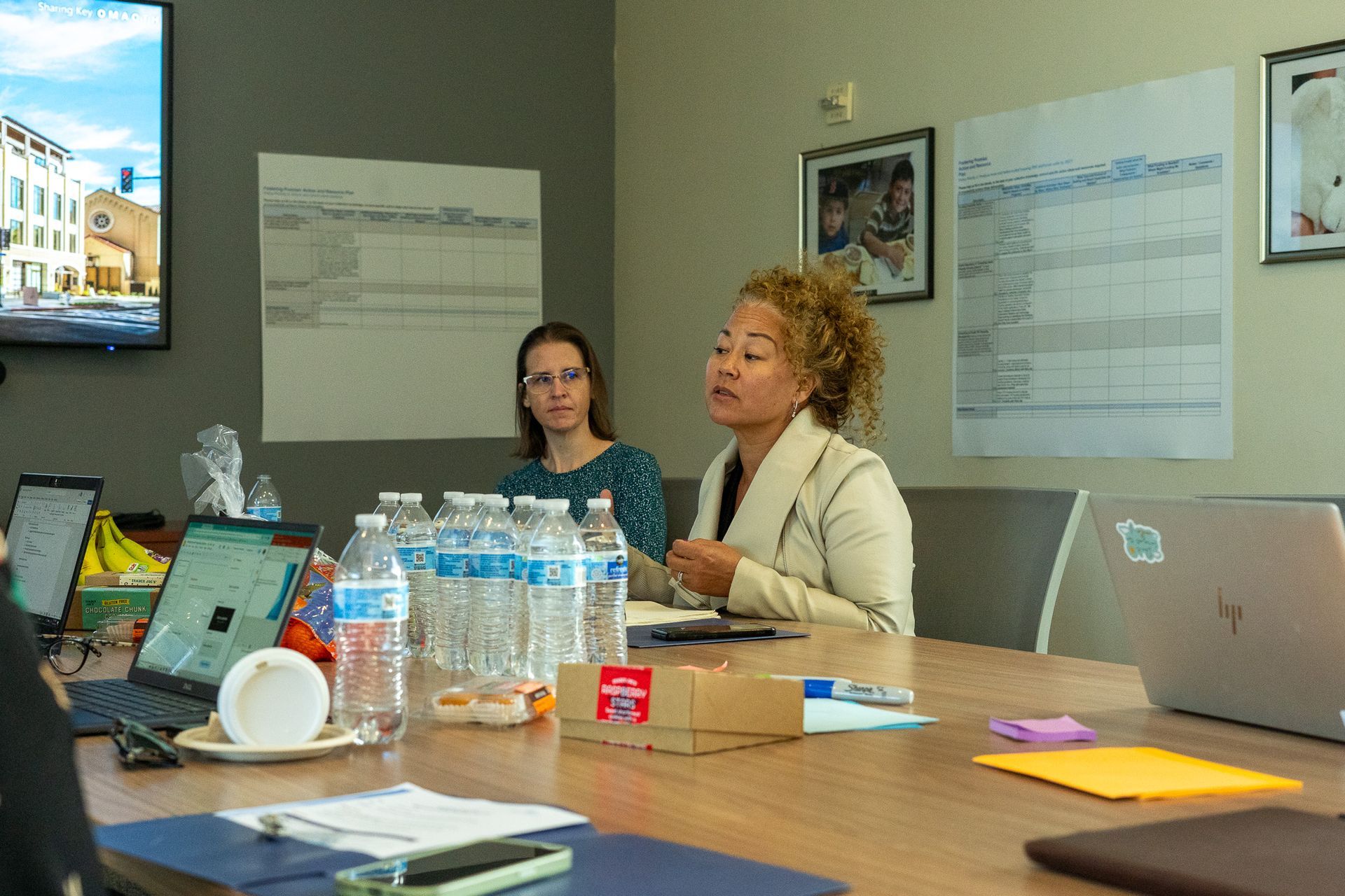 Two people seated at a conference table in a meeting room, with laptops, water bottles, and papers.