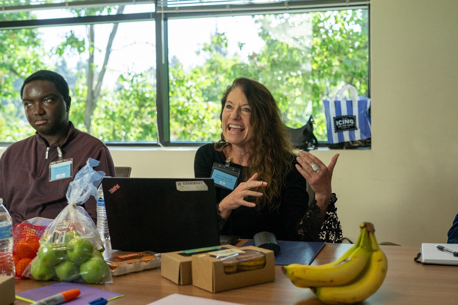 Two people seated at a table with fruit, a laptop, and papers in a bright room with large windows