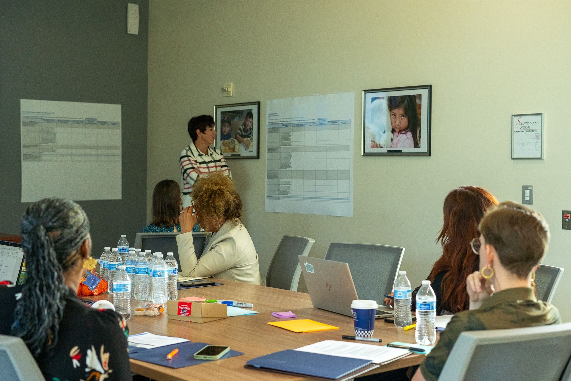 Small meeting room with several people seated around a conference table watching a presenter.