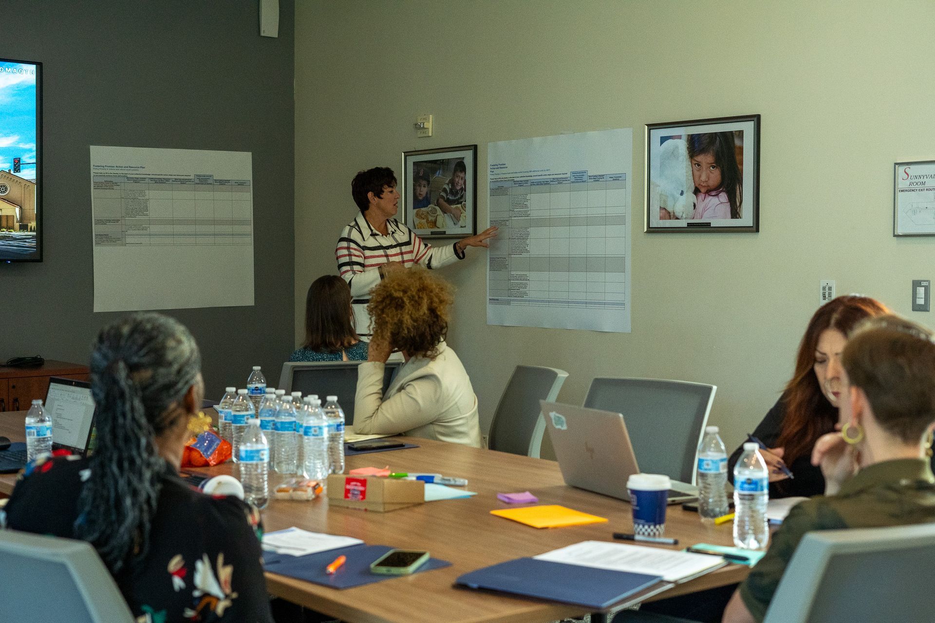 Small meeting room with people seated at a table while one person presents by a wall chart.