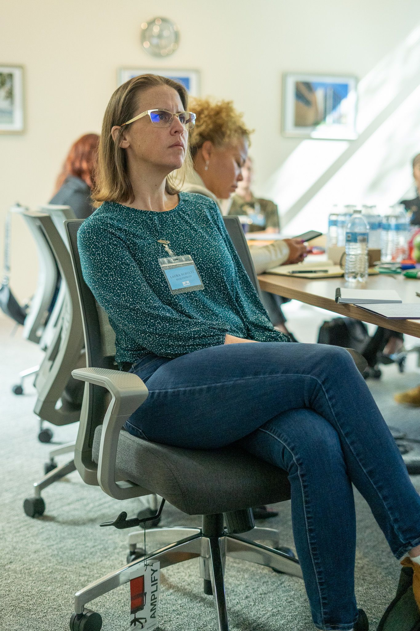 Woman in a teal patterned top sits in an office chair at a conference table, listening attentively.