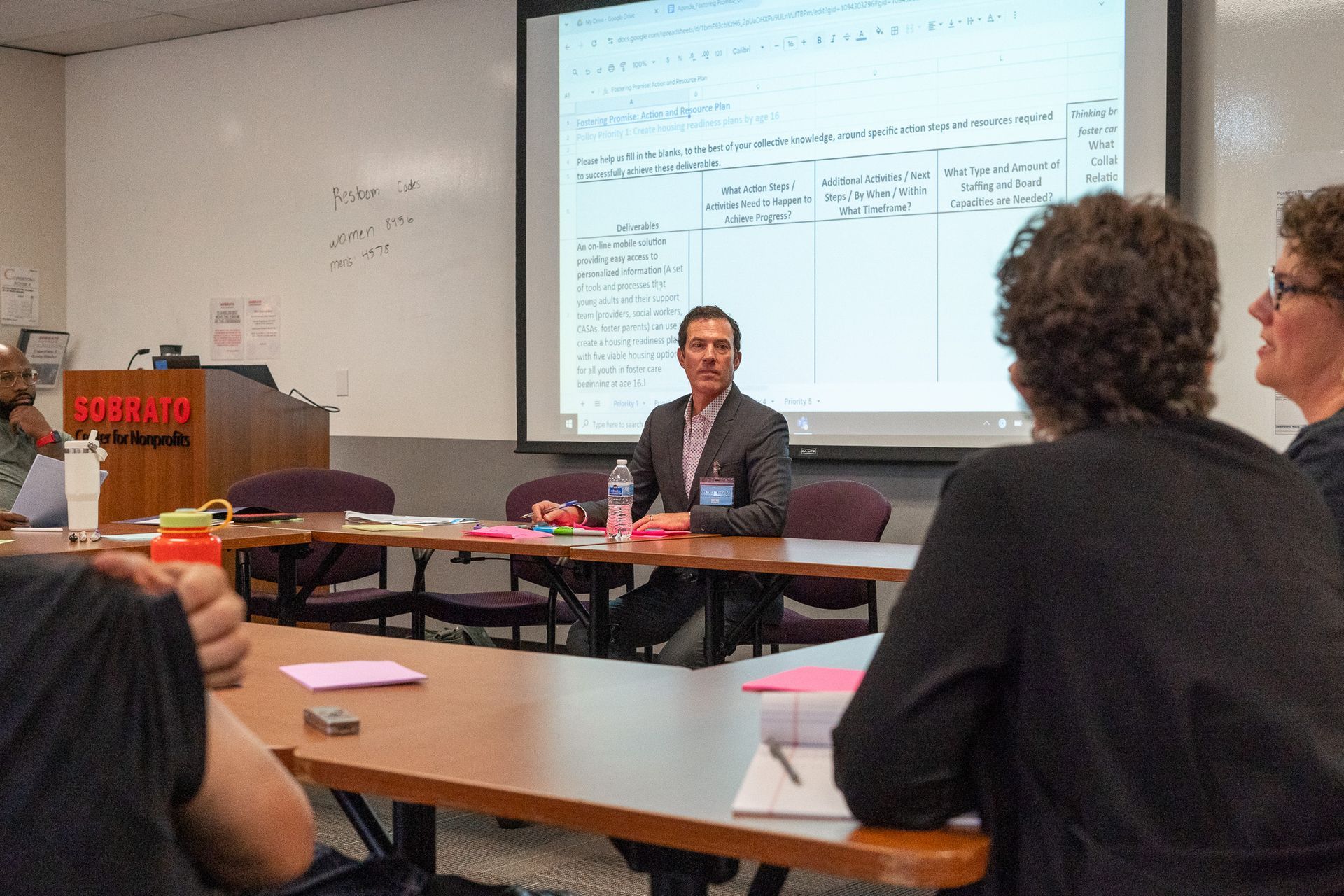 Classroom lecture with a speaker at the front and students seated at tables facing a projected presentation.