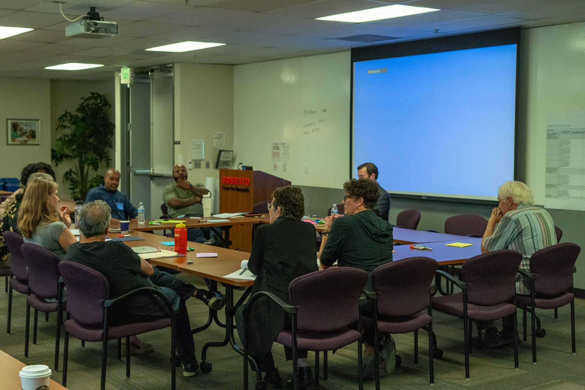 People seated around tables in a conference room facing a presentation screen.