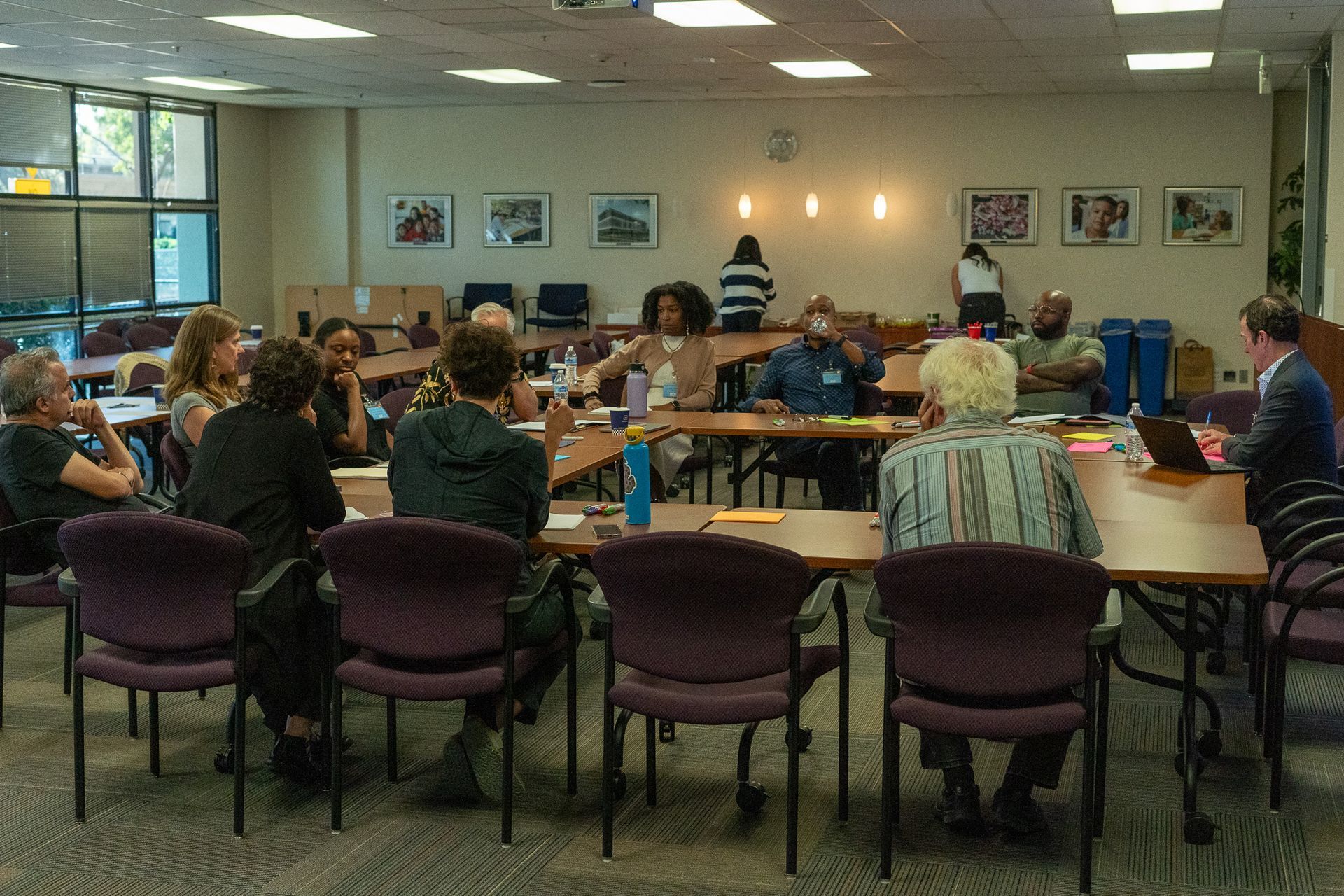 People seated around tables in a meeting room, with framed pictures on the wall and fluorescent lighting.