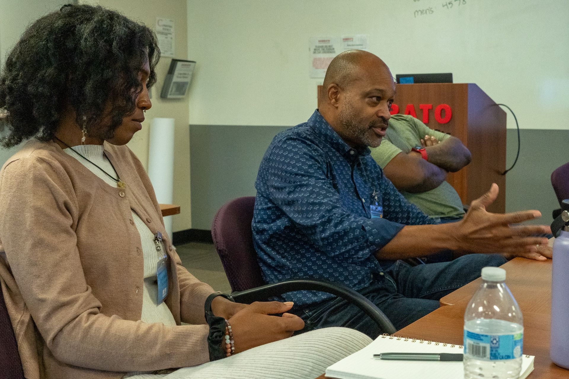 Two people sit in a meeting room, talking across a table with a laptop, papers, and a water bottle.