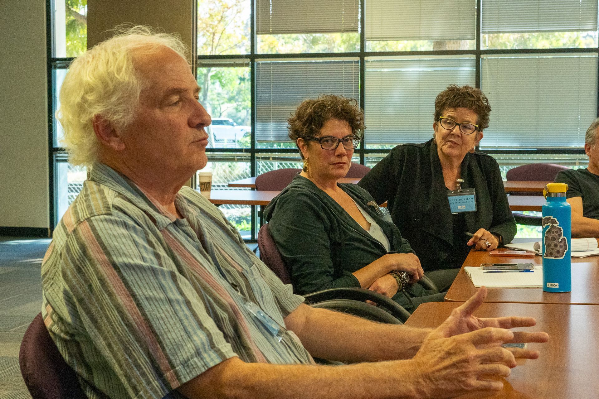 Three people sit at a table in a bright meeting room, talking with papers and a water bottle nearby.