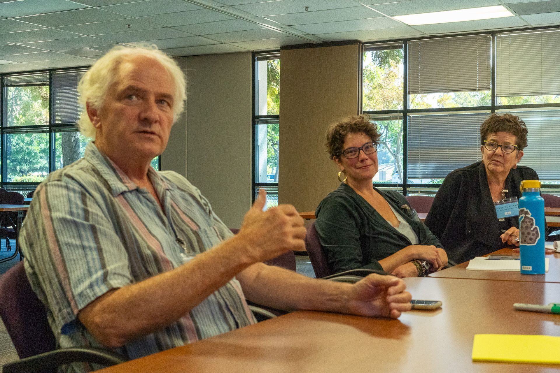 Three people seated at a conference table in a bright meeting room, one giving a thumbs-up