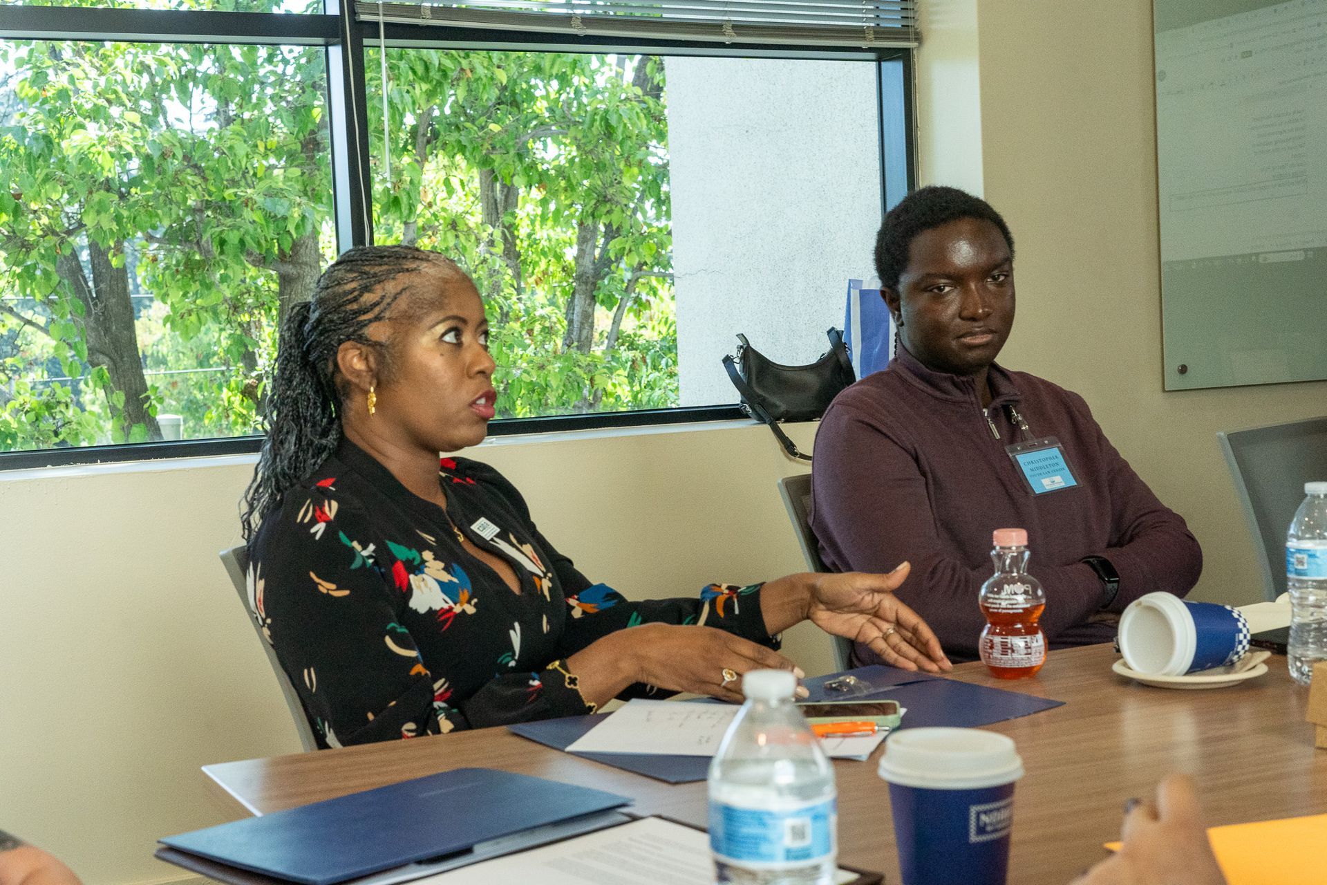 Two people seated at a conference table in a meeting room, with notebooks and water bottles nearby.