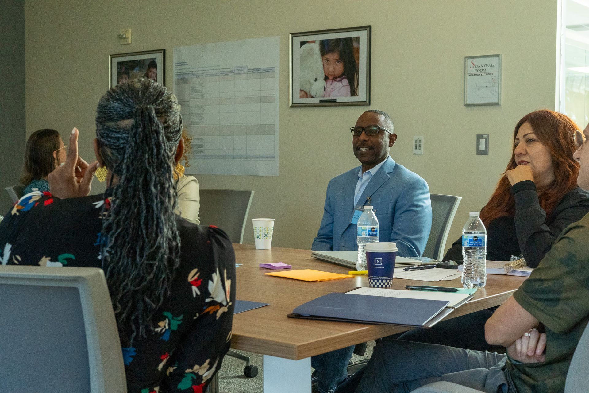 People seated around a conference table in a meeting room, with papers, laptops, and a framed photo on the wall.