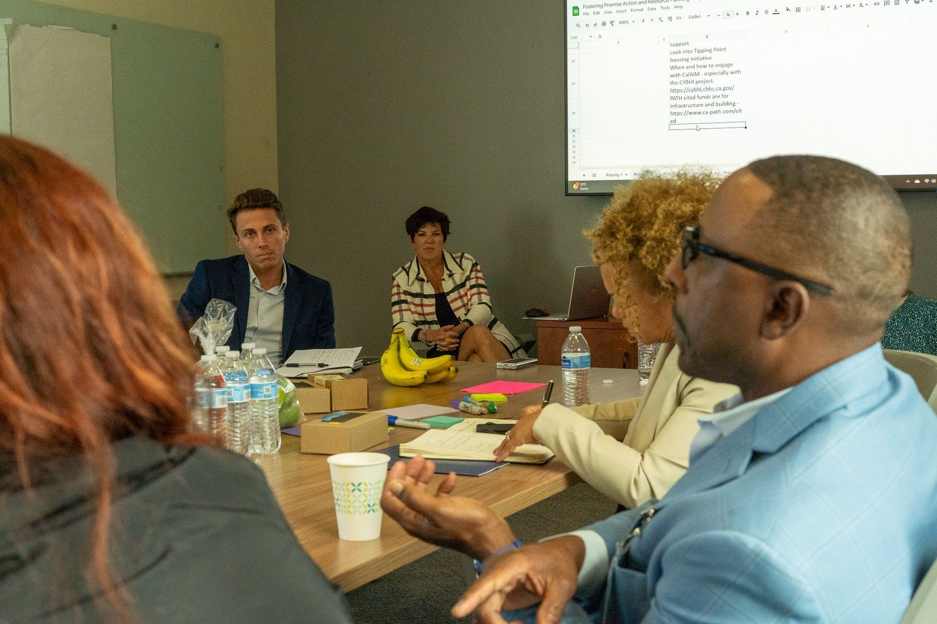 People in a meeting room gathered around a conference table, with laptops, papers, and a projected screen.