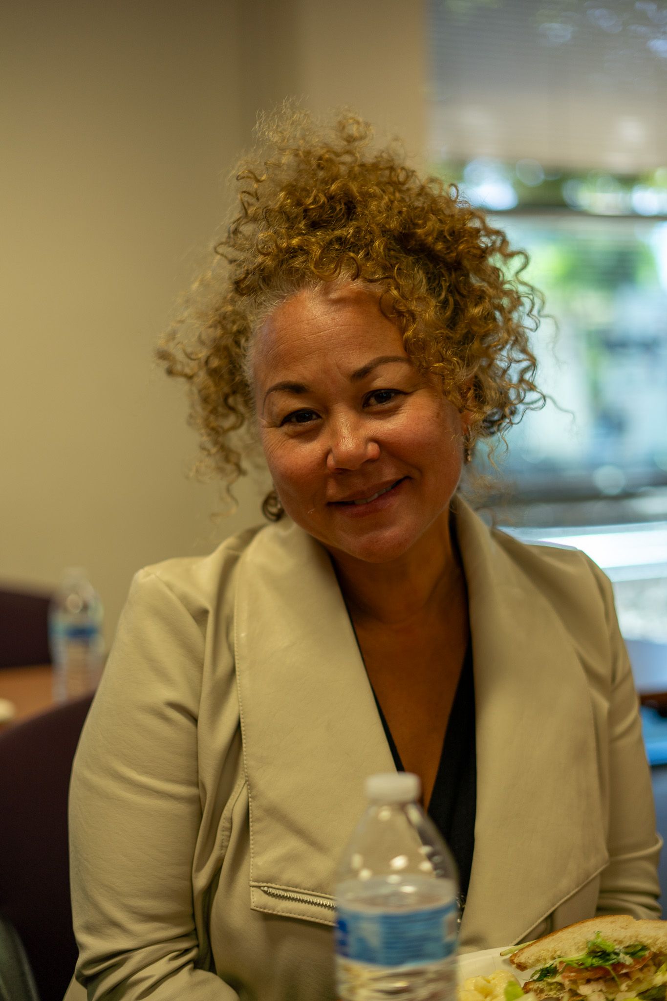 Smiling woman in a cream blazer seated indoors with a water bottle in front of her