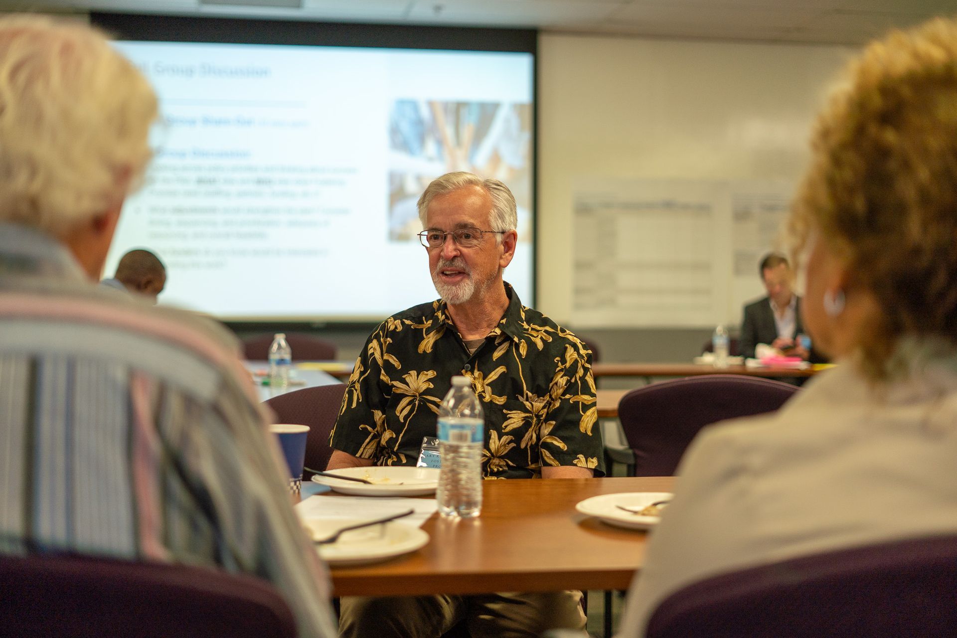 Meeting attendees seated around a conference table with a speaker in front of a presentation screen