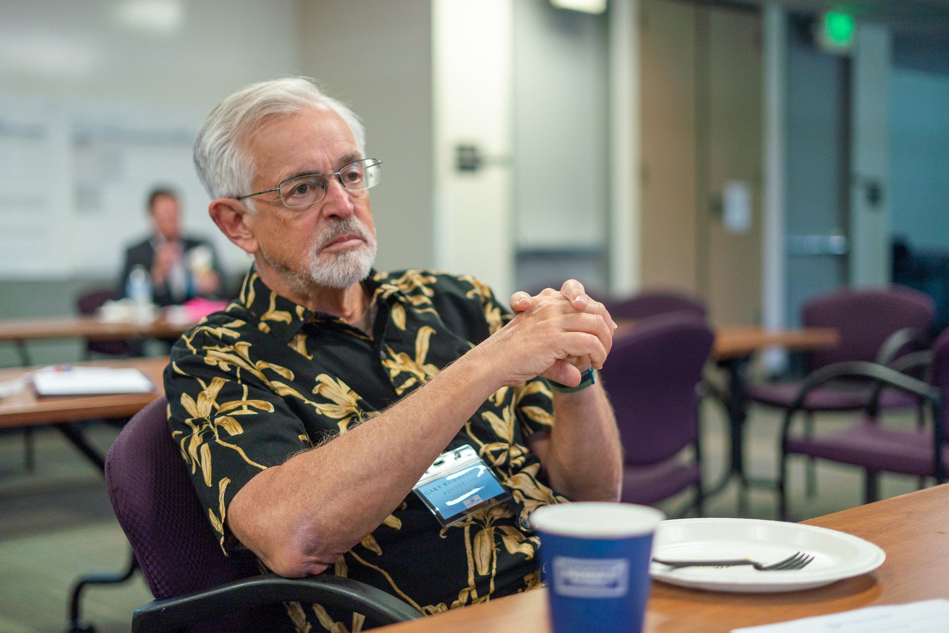 Man seated at a table in a conference room, hands clasped, with a coffee cup in the foreground.
