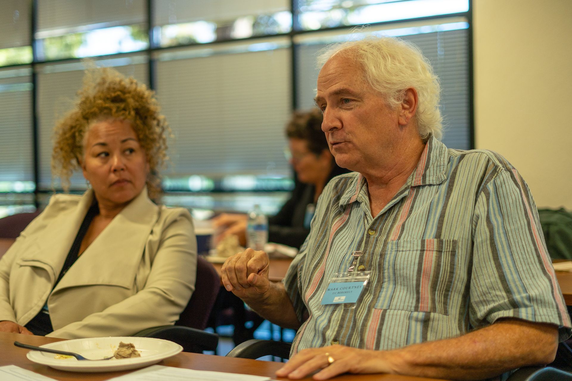 Two people seated at a table, talking in a bright indoor setting with windows behind them.