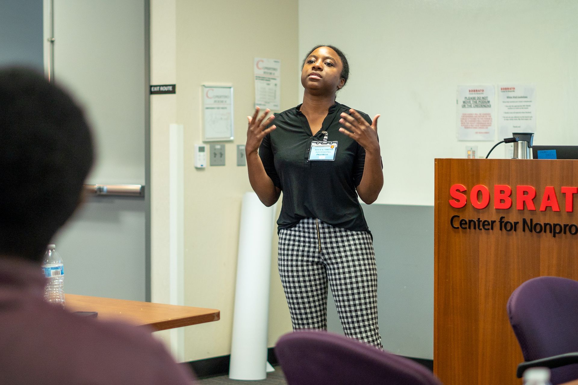 Presenter speaking at a podium in a conference room, with a sign reading 