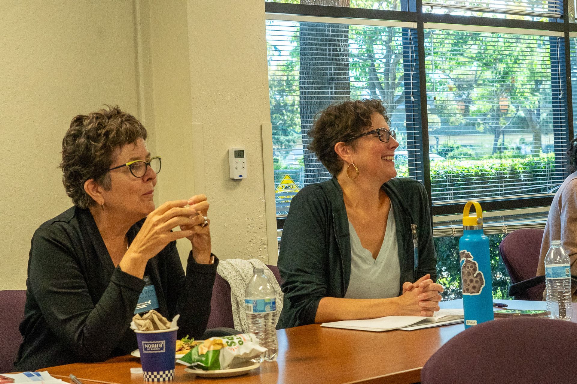 Two people seated at a table in a meeting room, listening and taking notes.