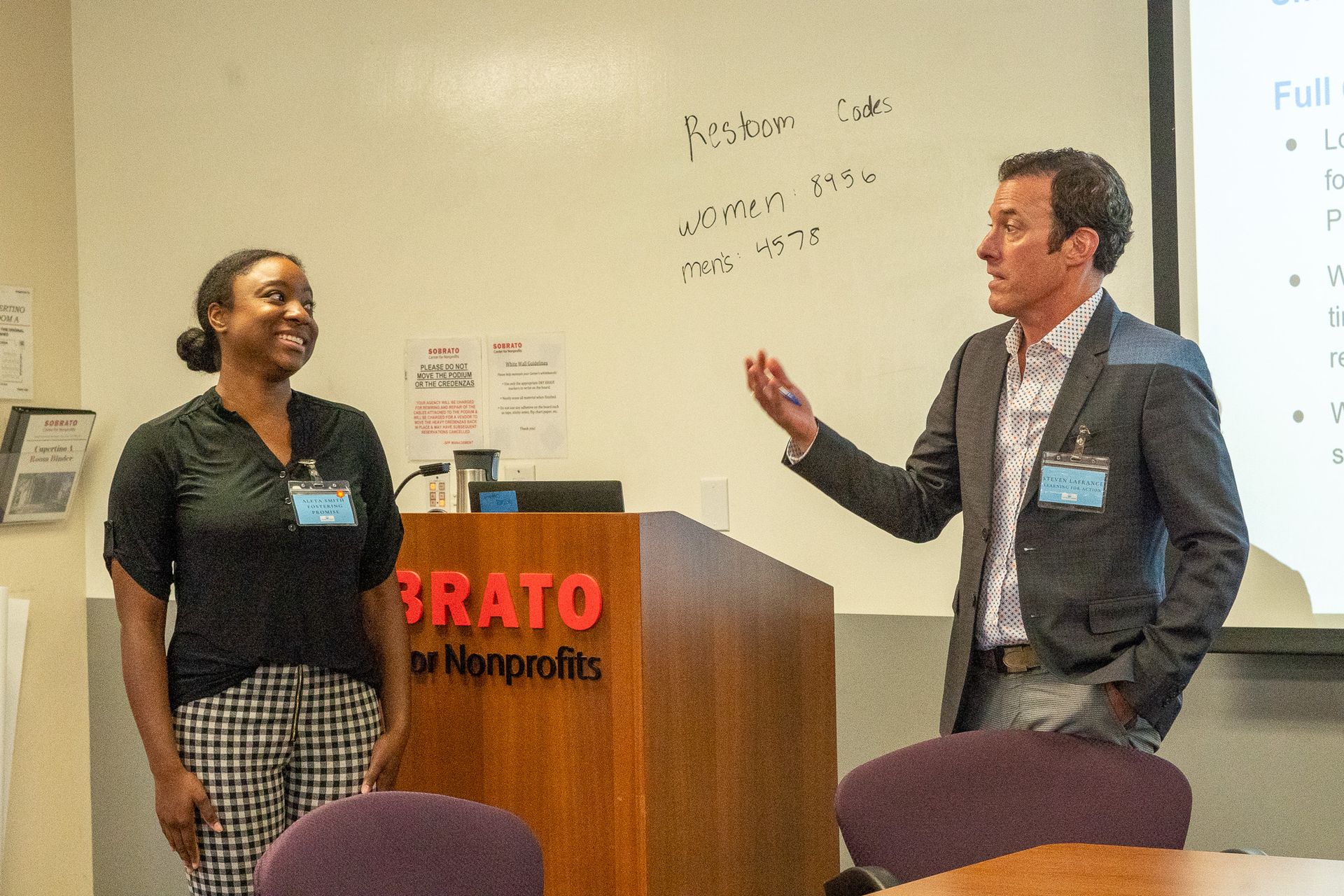 Two people speaking beside a podium in a classroom or conference room, one gesturing toward a projection screen.