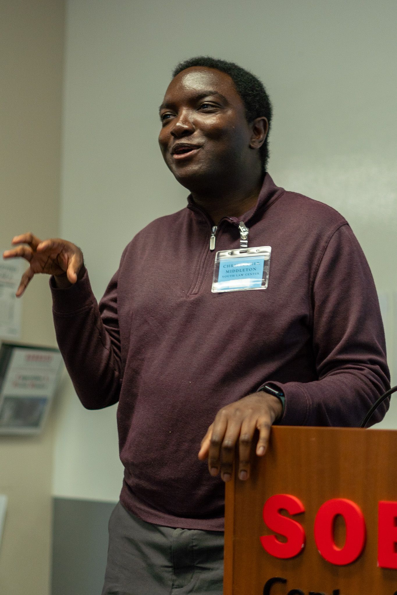 Person speaking at a podium, wearing a maroon sweater and name badge, gesturing with one hand.