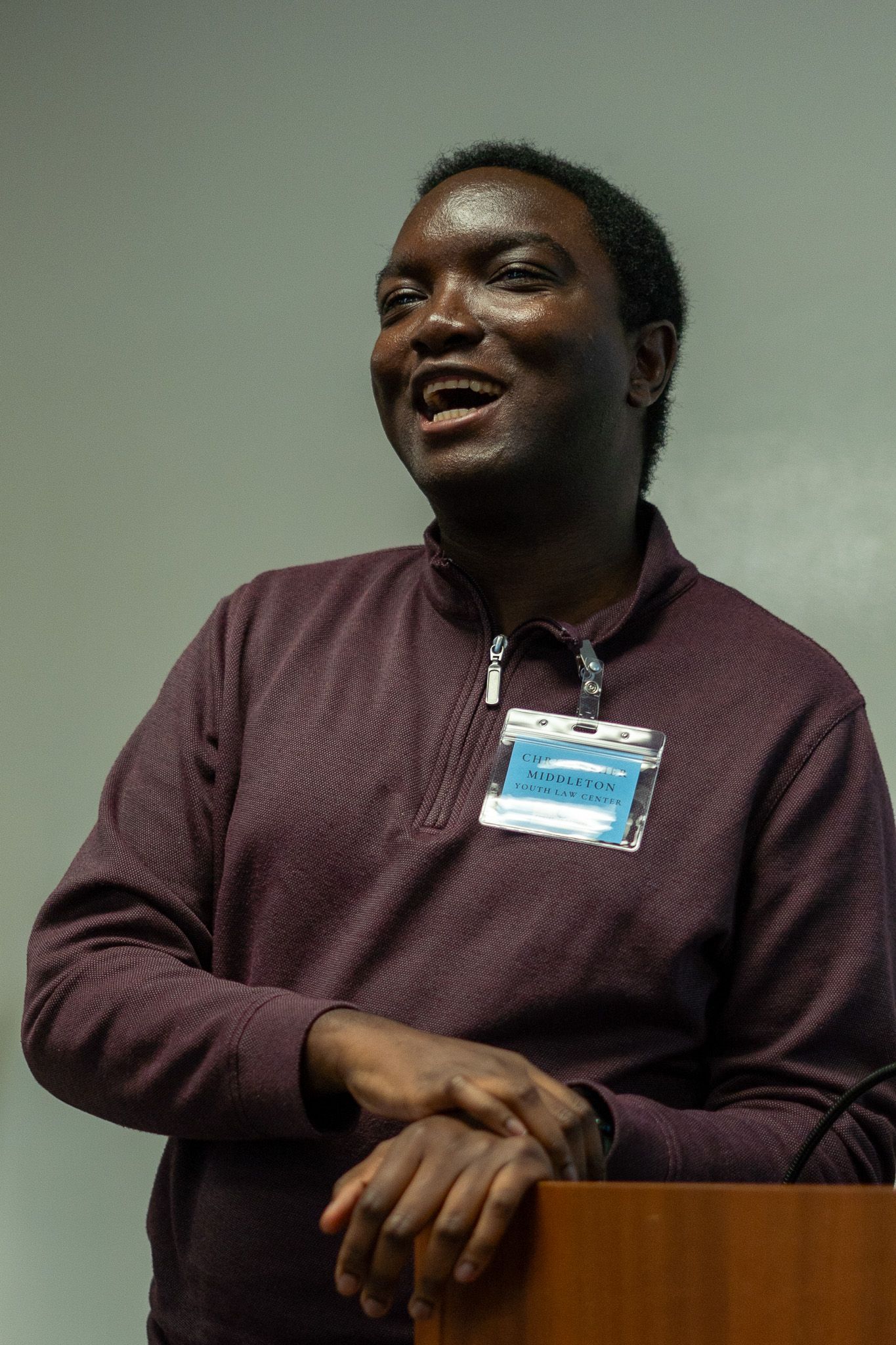 Man standing behind a wooden podium, wearing a purple sweater and name badge, speaking in front of a plain wall