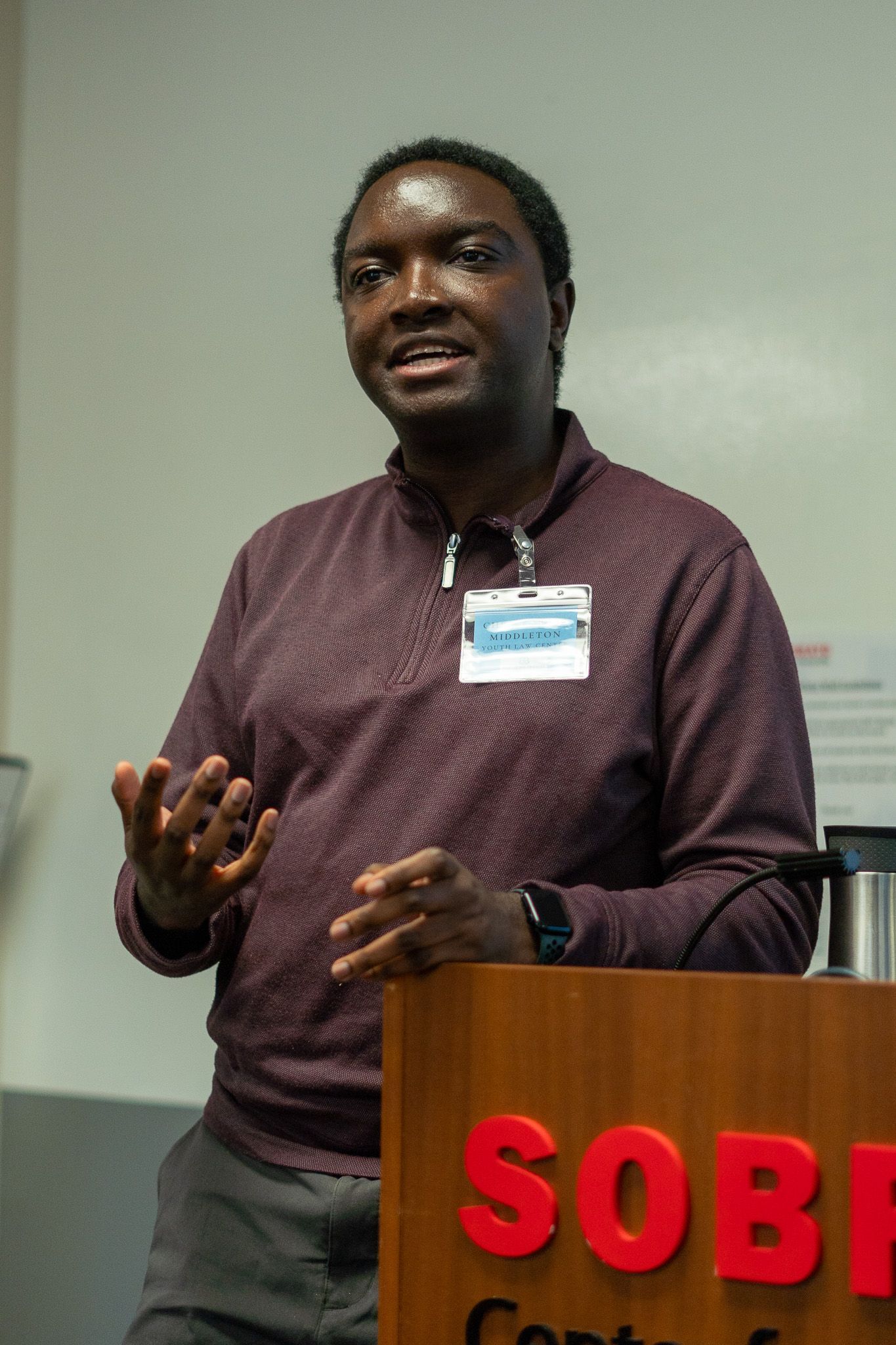 Man speaking at a podium with a red “SOBE” sign, gesturing in a classroom or conference room.