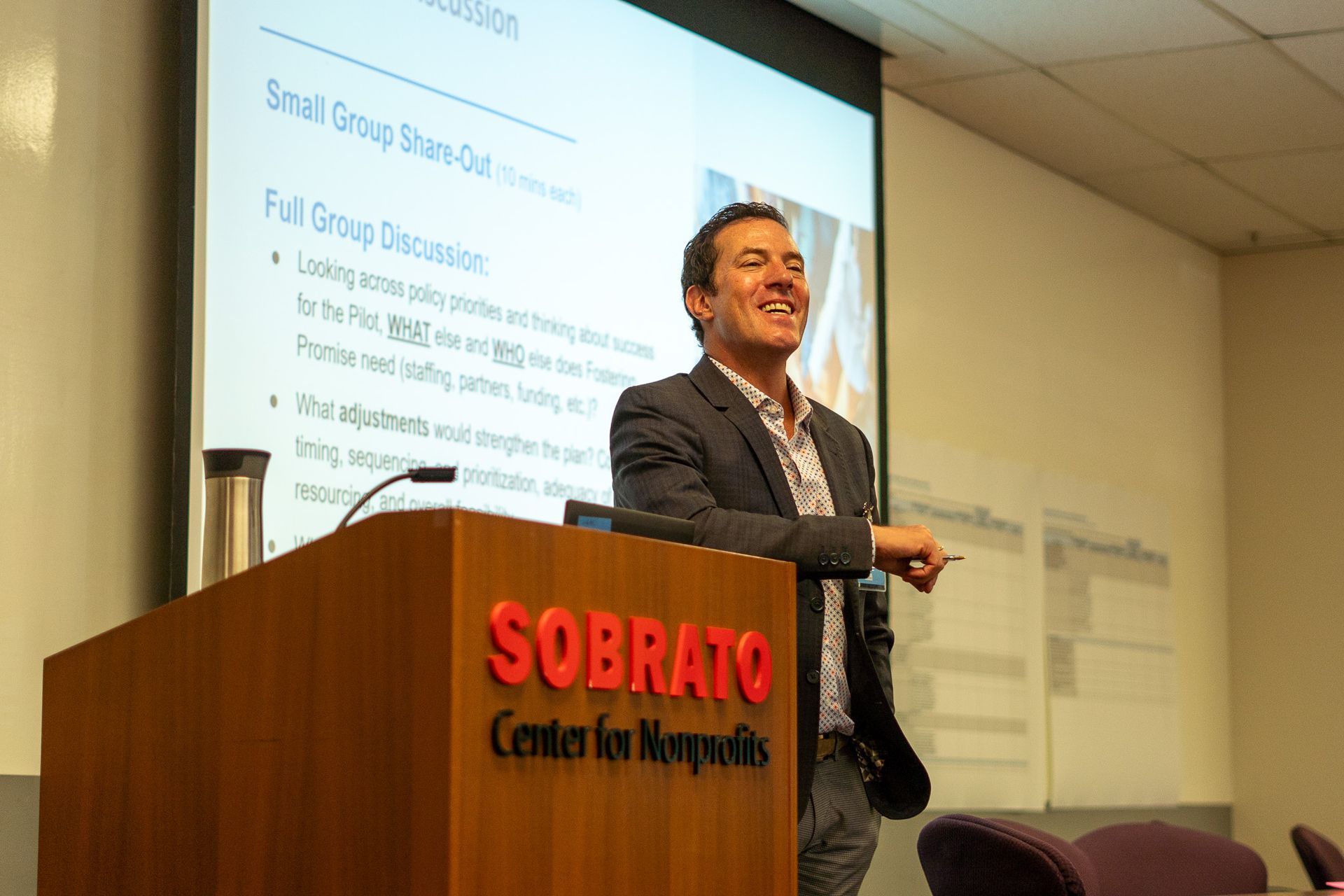 Man speaking at a podium in a conference room, with a presentation slide projected behind him.