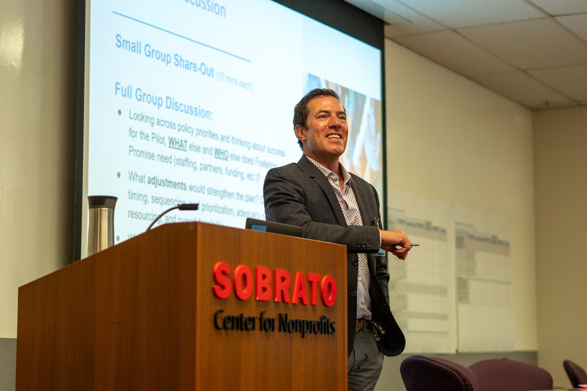 Man speaking at podium in conference room, with presentation slide projected behind him