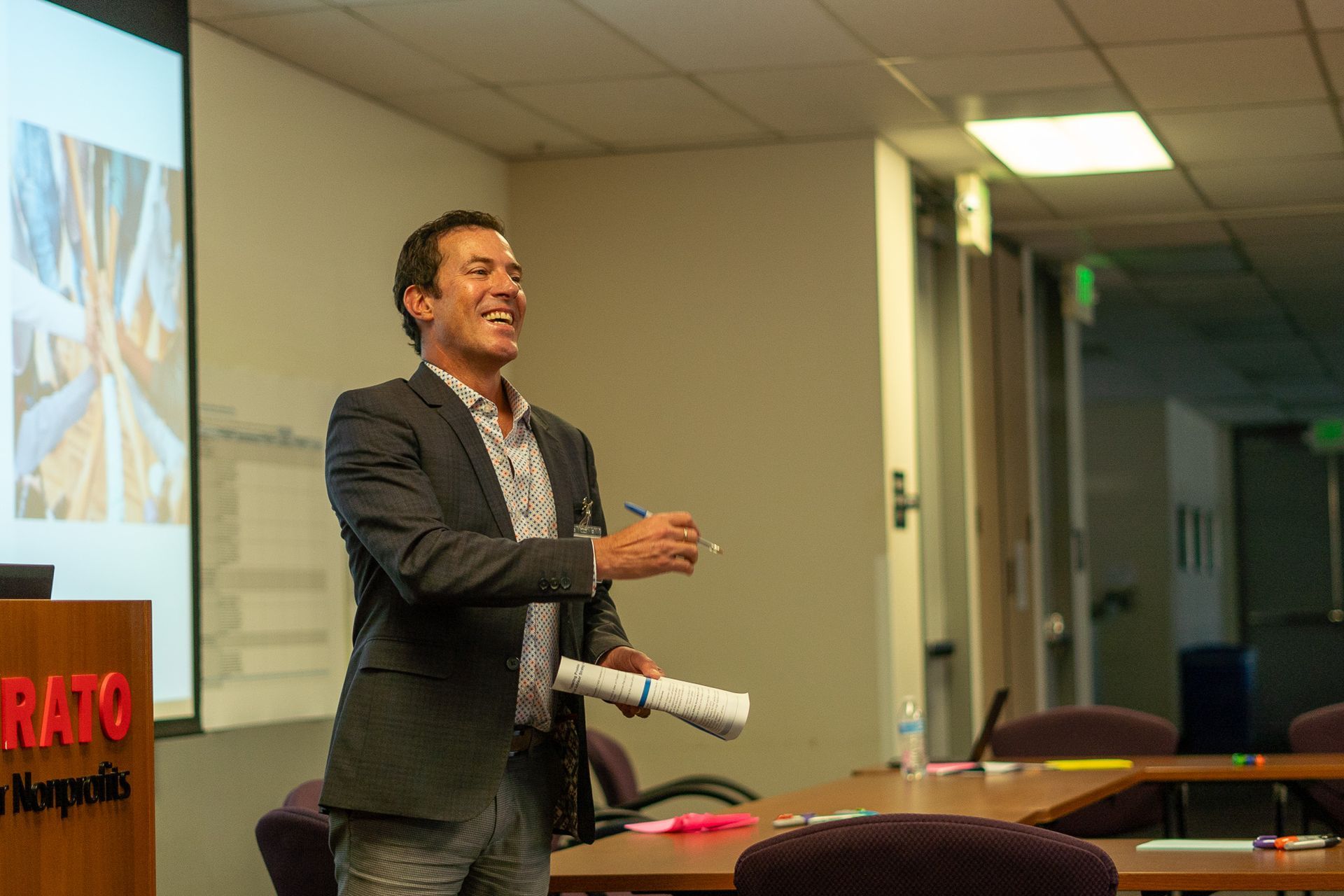 Man in a blazer speaking in a conference room, gesturing beside a projection screen and table