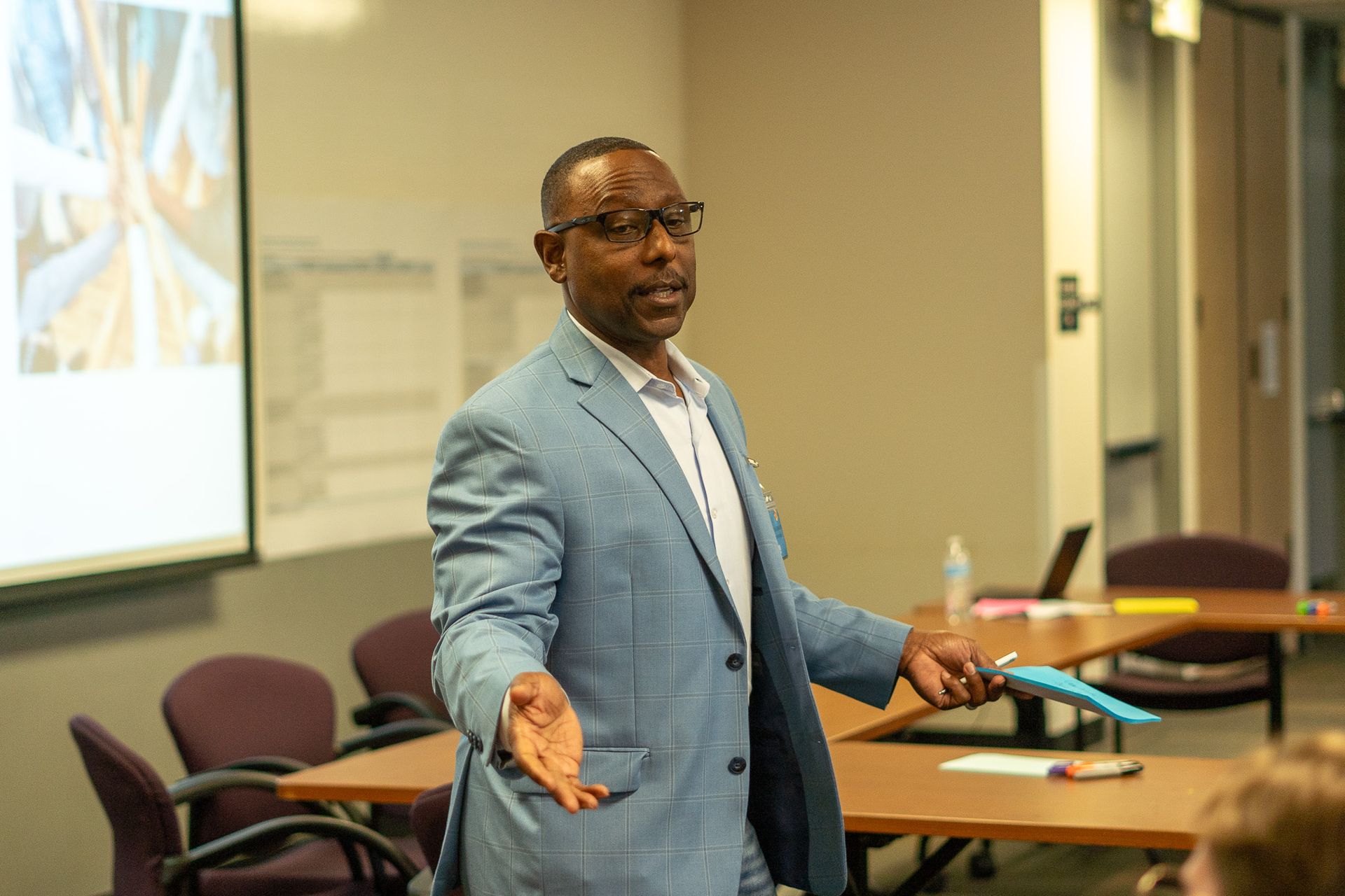 Man in a blue blazer speaking in a conference room, gesturing with open hands at a table.