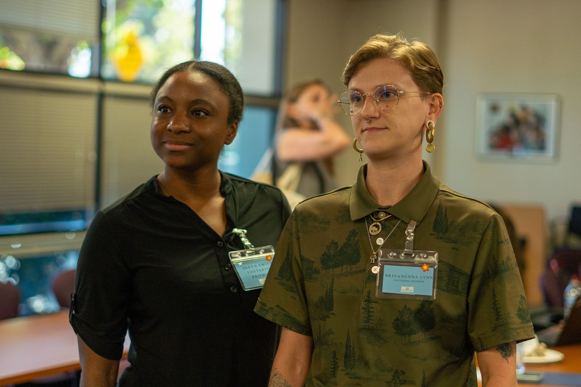 Two staff members standing indoors, one in a black shirt and one in a camouflage uniform with glasses and a name badge.