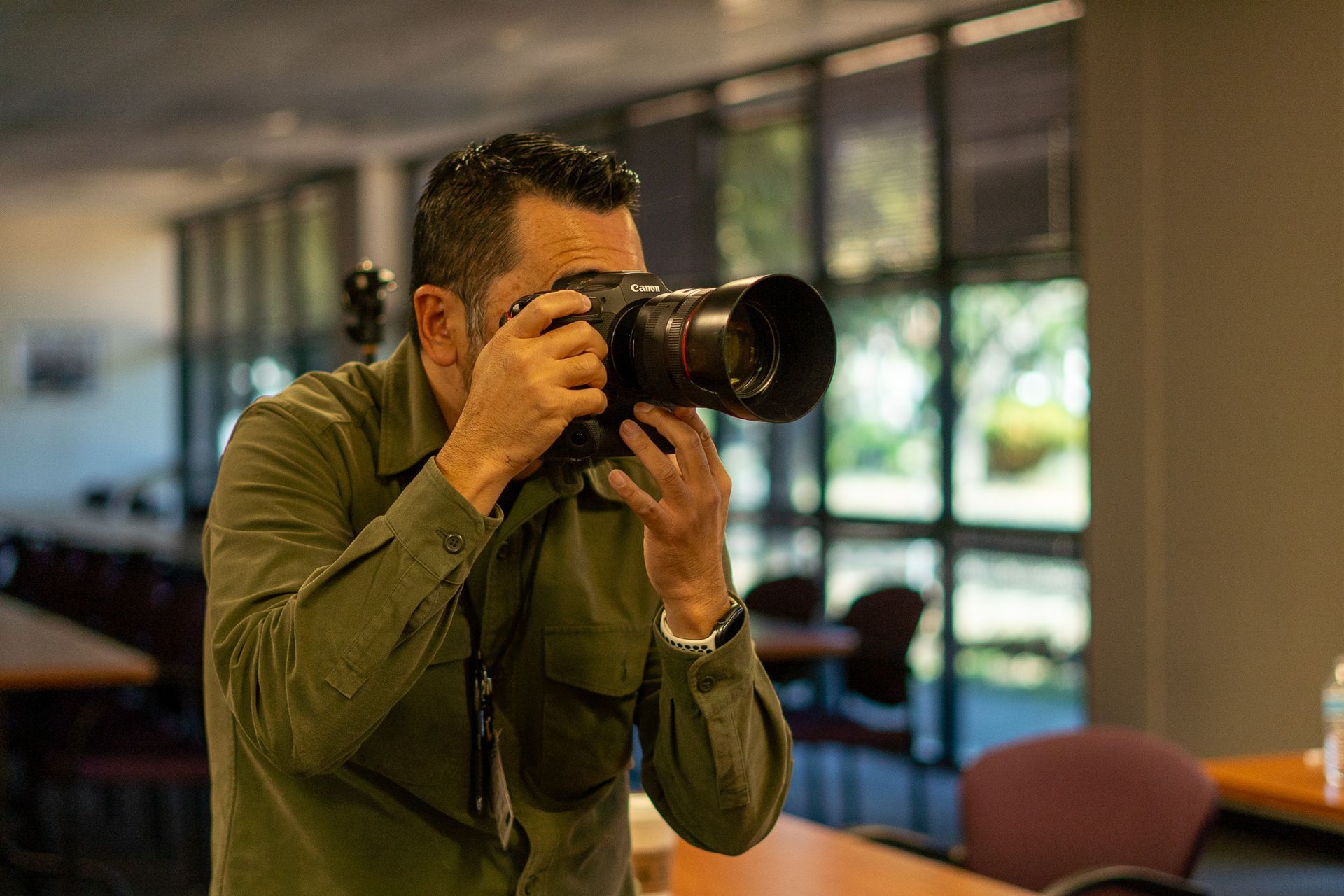 Photographer in an olive shirt aims a large camera indoors near windows and tables.