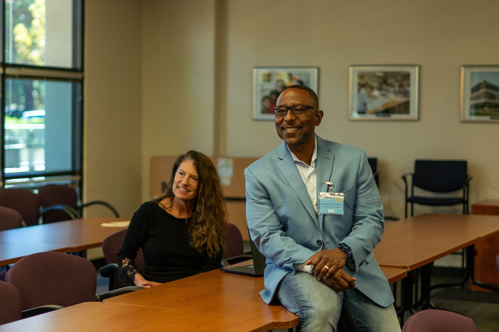 Two people sitting at a conference table in a meeting room, smiling at the camera.