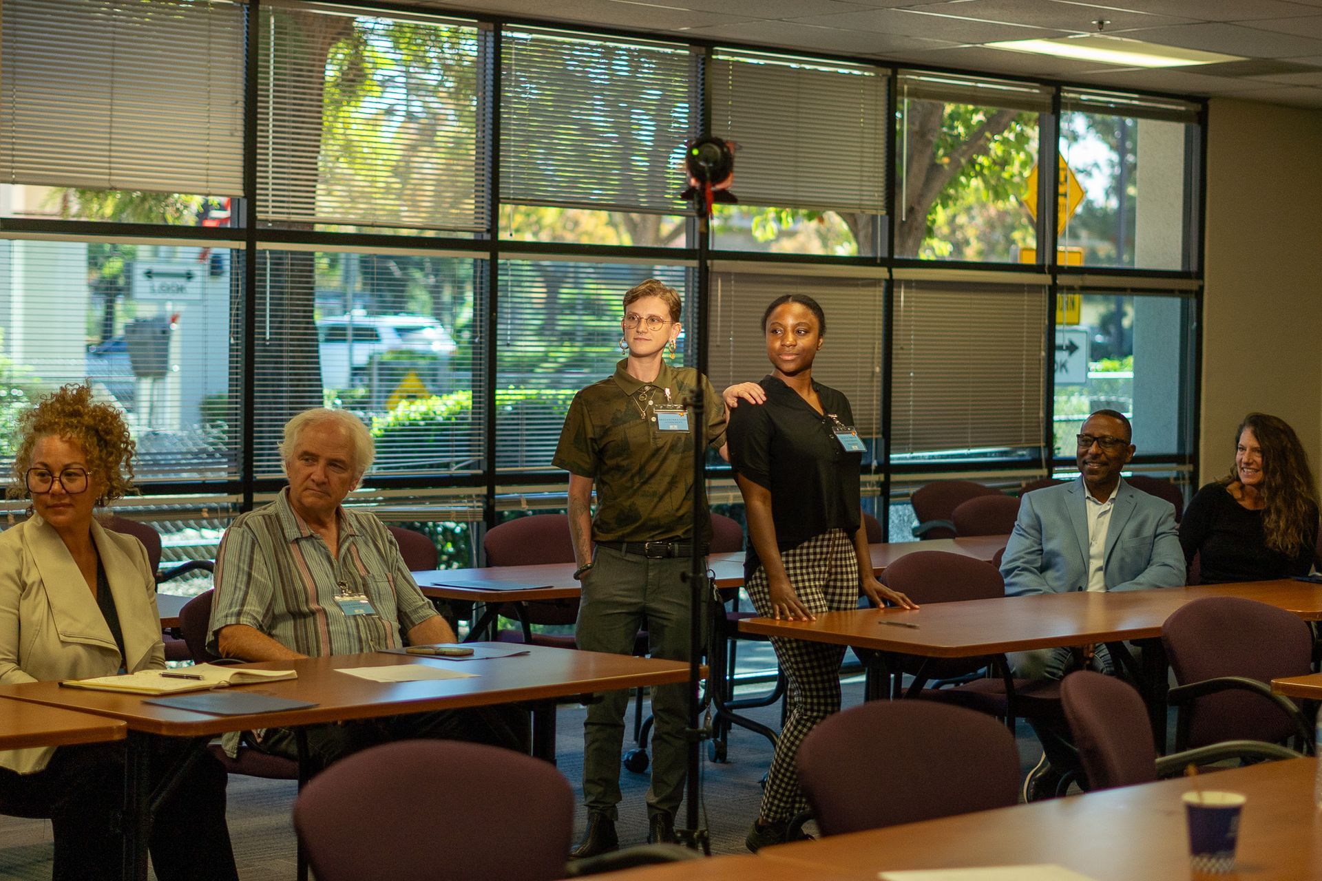 People seated at tables in a sunlit meeting room with large windows and potted plants