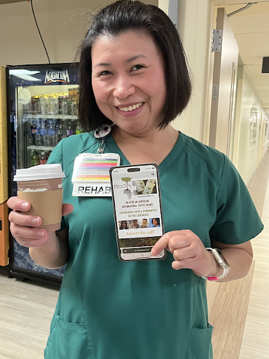 Smiling hospital worker in green scrubs holding coffee and a phone, standing in a hallway