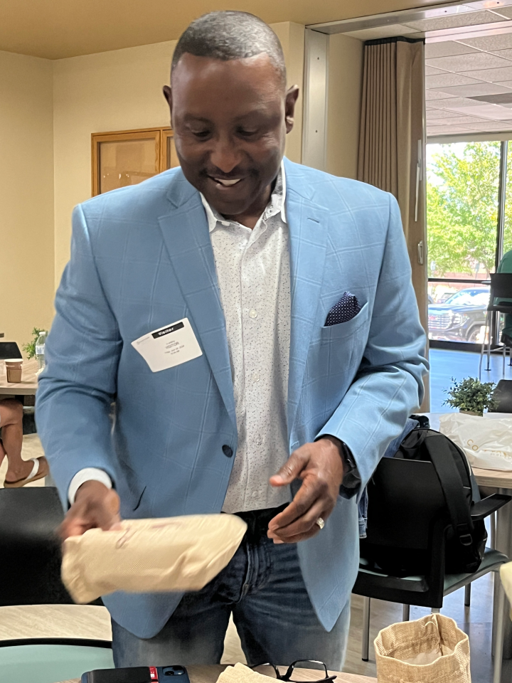 Man in a light blue blazer smiling while holding a beige wrapped item in a room with tables and chairs