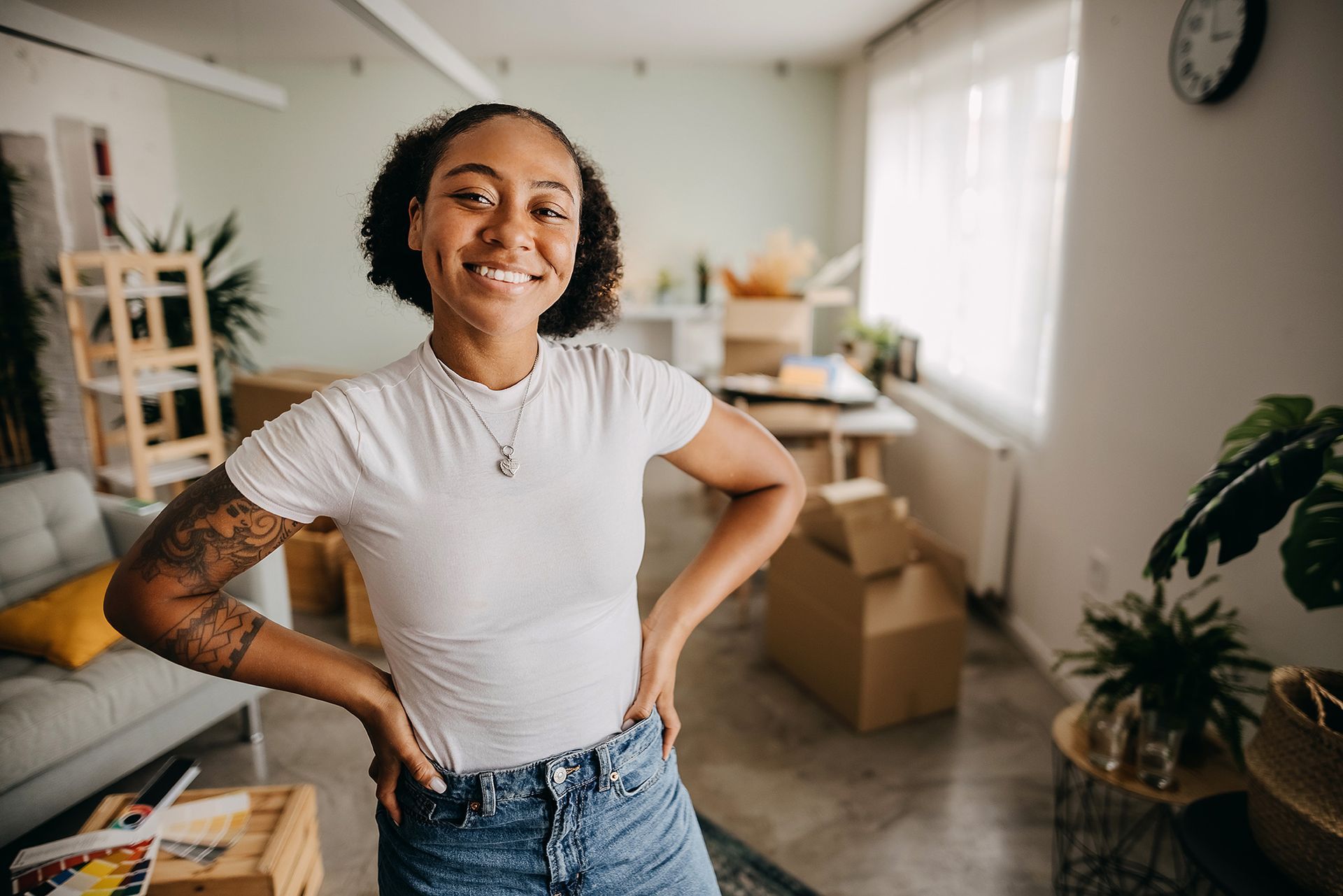 Person smiling with hands on hips in a bright room with moving boxes and plants