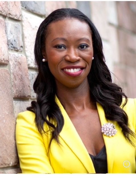 Smiling woman in a yellow blazer standing against a stone wall.