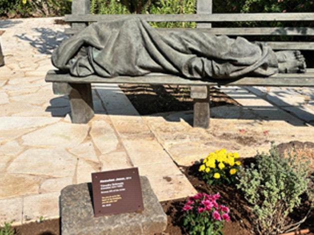 Stone sculpture of a sleeping figure on a bench beside a plaque and yellow and pink flowers outdoors