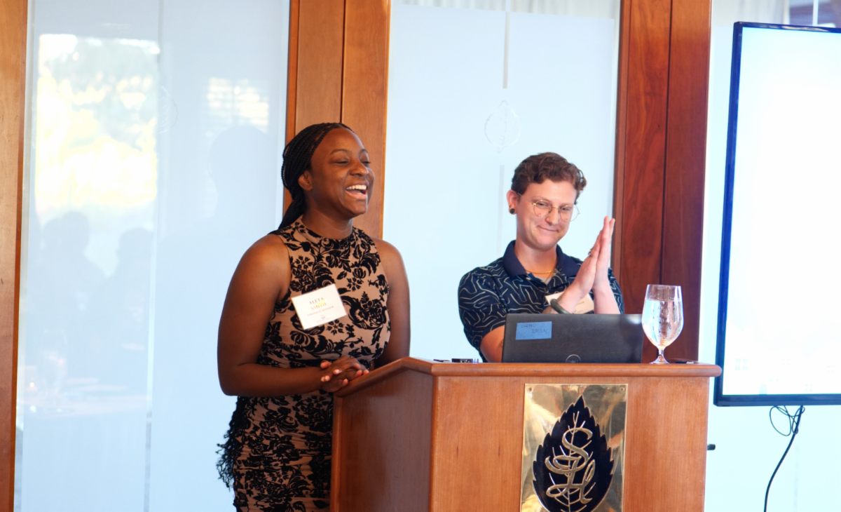 Two speakers at a podium during a presentation, one speaking and one clapping beside a laptop and screen.