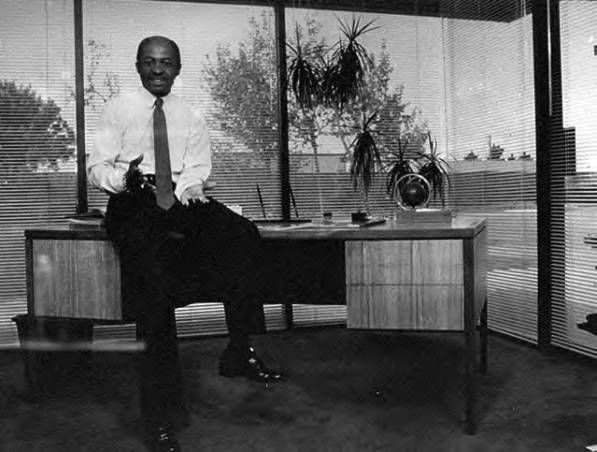 Man seated on a desk in an office, with large windows and a potted plant in the background.