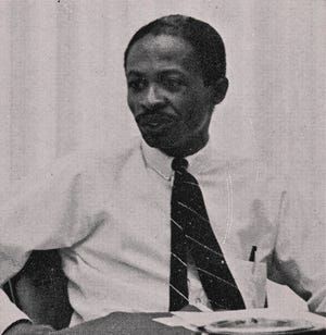 Man seated at a table, wearing a white shirt and dark striped tie, looking to the side.